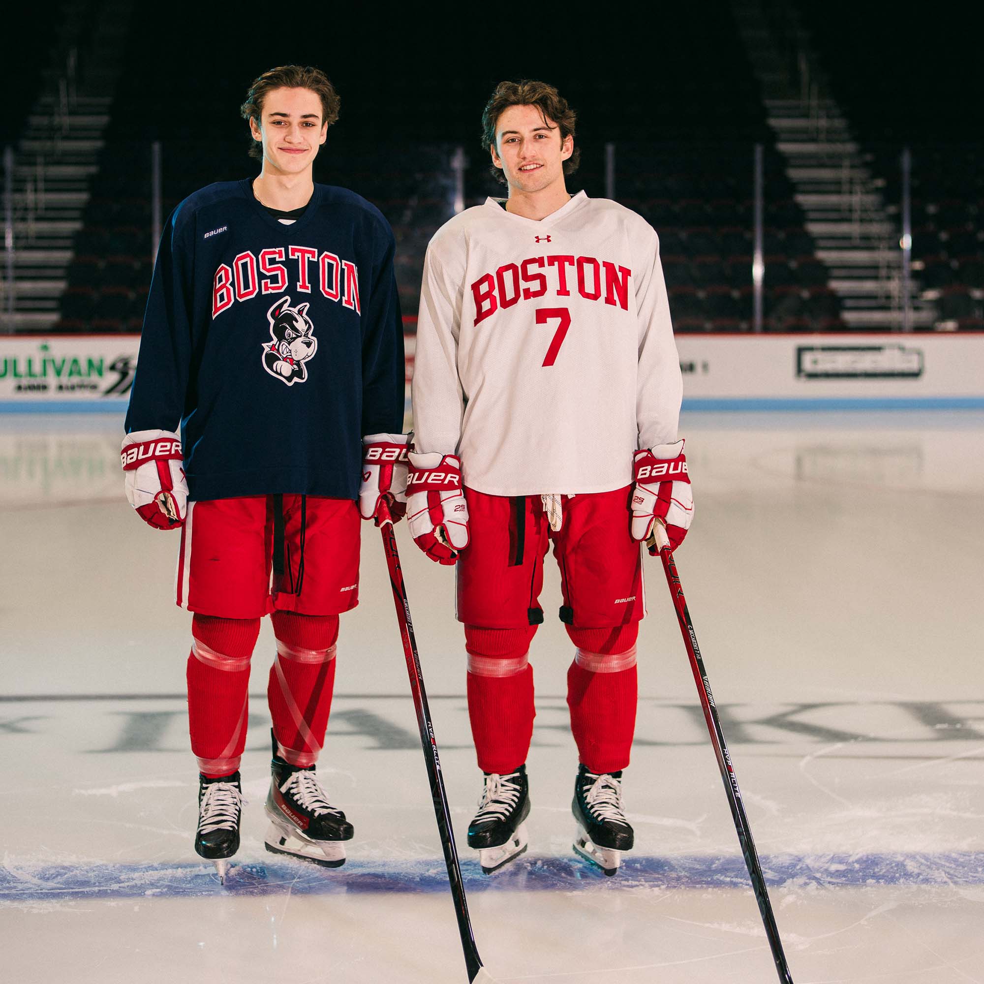 Photo: Two hockey players pose standing on the ice facing the camera. One wears a black practice jersey and the other, a white practice jersey and the red issue hockey pants.