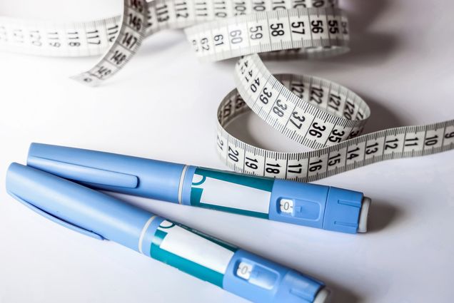 Photo: Two Ozempic Insulin injection pens laid across one another with a measuring tape all rolled out behind the pens. They lay on a white surface.