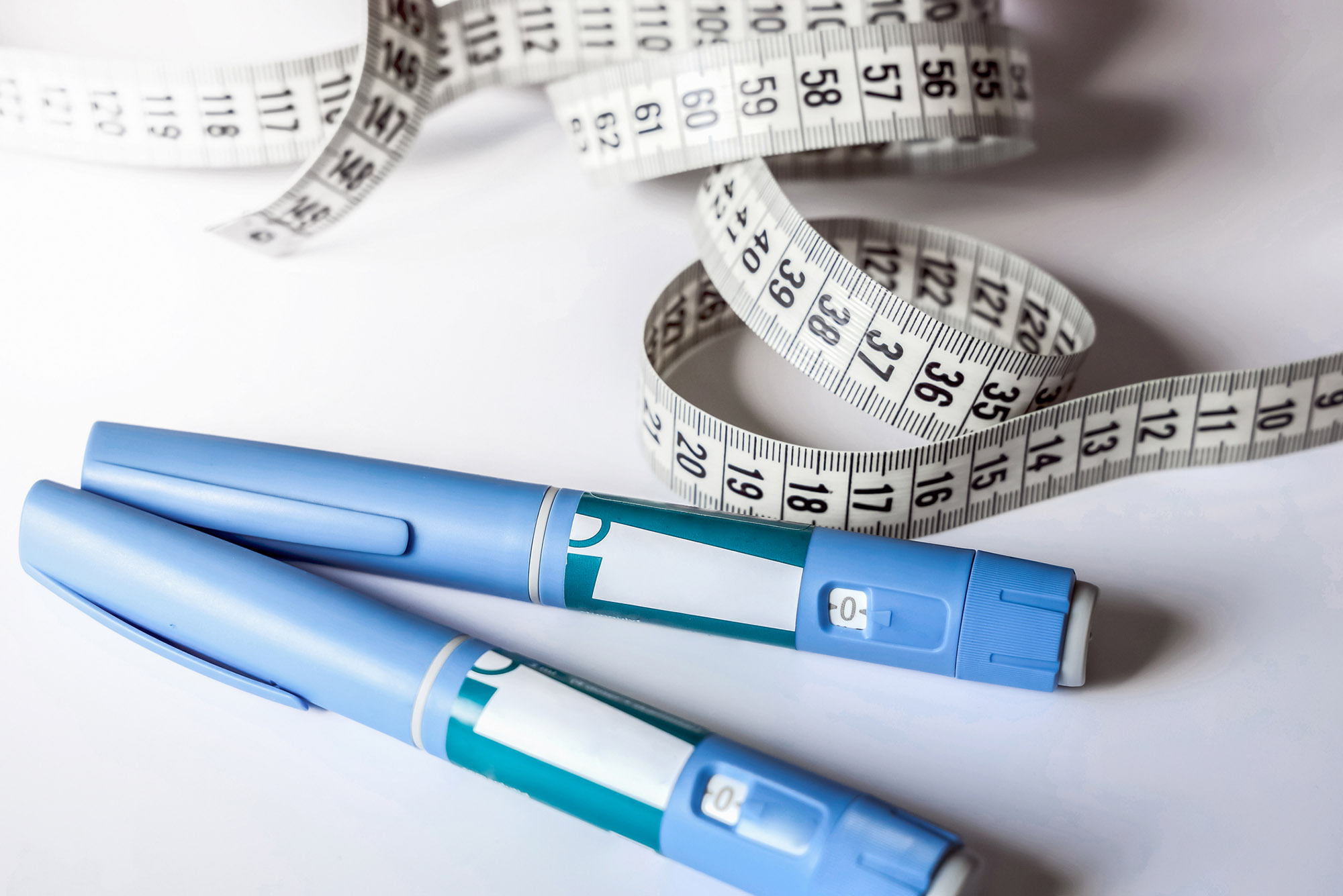 Photo: Two Ozempic Insulin injection pens laid across one another with a measuring tape all rolled out behind the pens. They lay on a white surface.