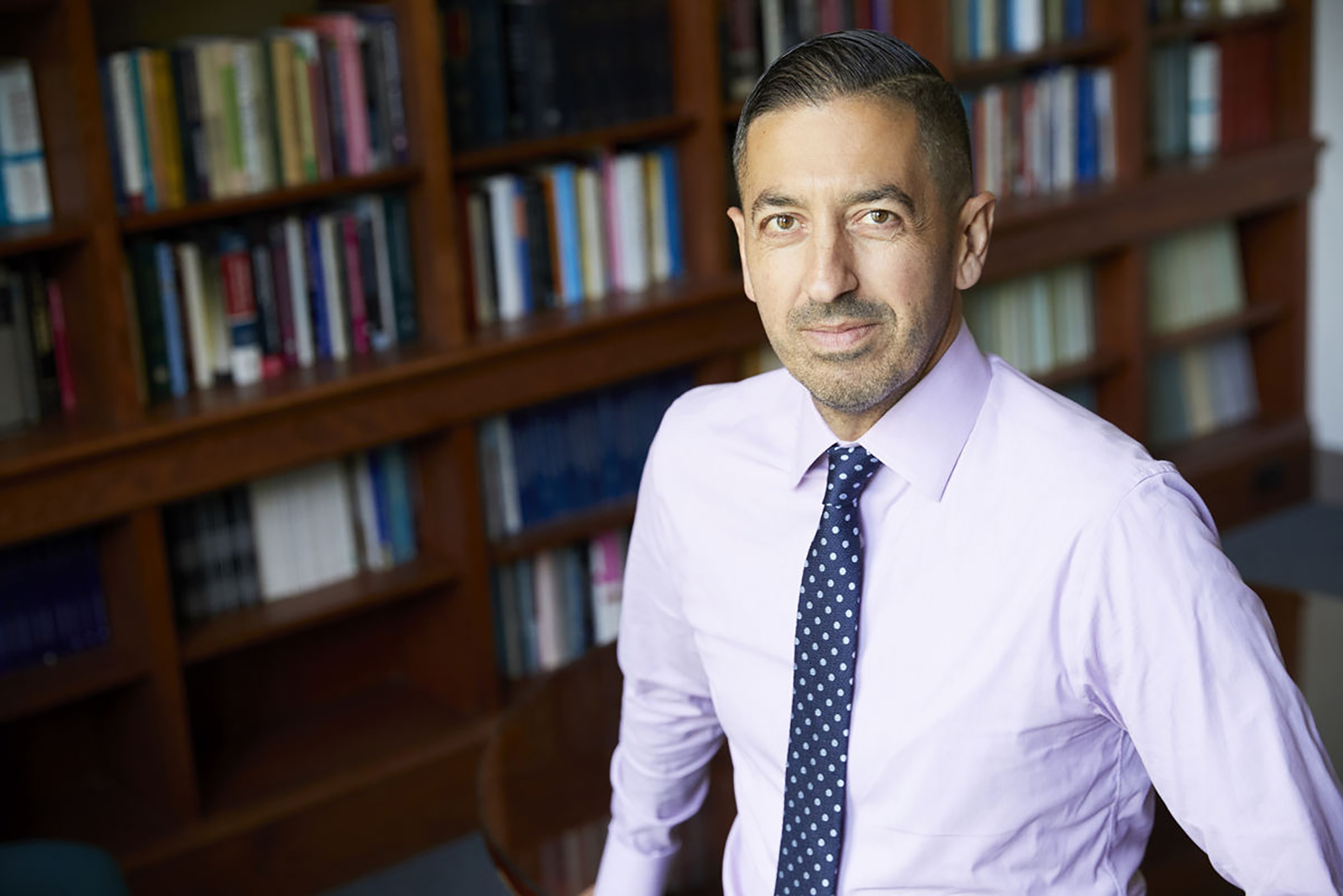 Photo: Sandro Galea, a man with stubble, wears a white button up with a dark tie in an office-setting with books lining the wall behind him. He poses for the photo with a serious face.