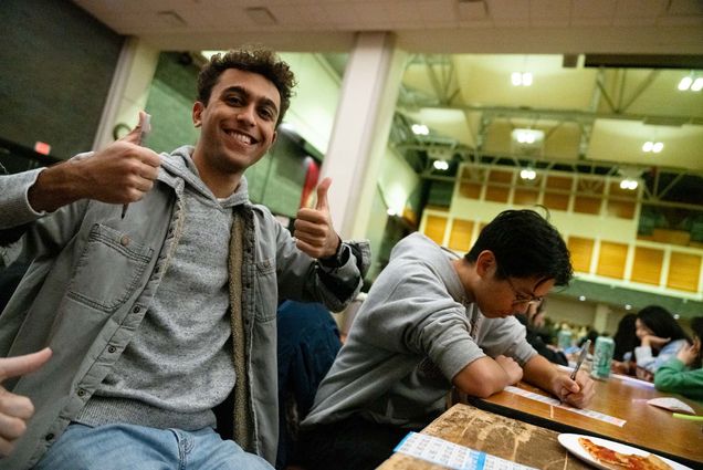 Photo: A young man holds two thumbs up while the man next to him fills in a bingo card.