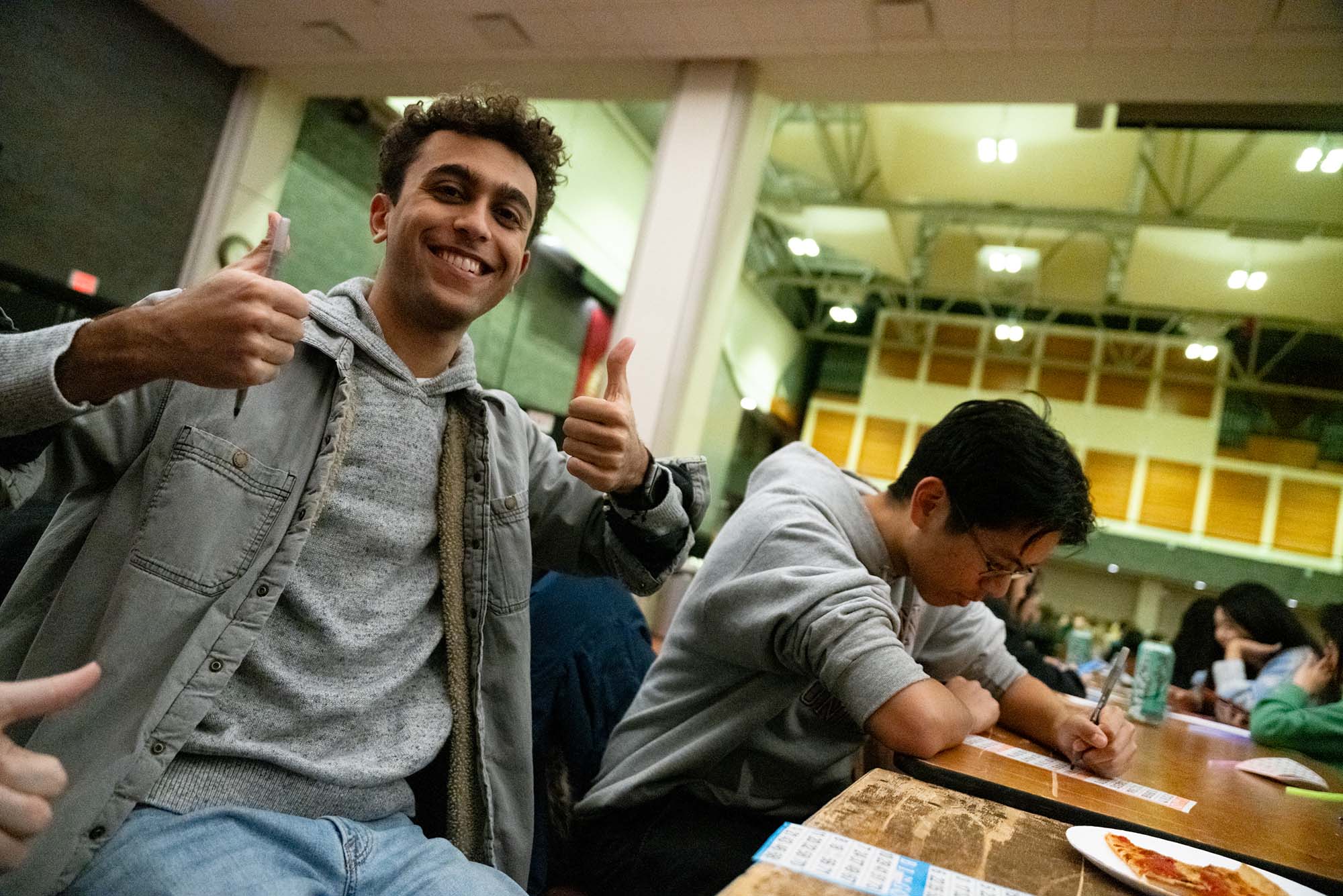 Photo: A young man holds two thumbs up while the man next to him fills in a bingo card.