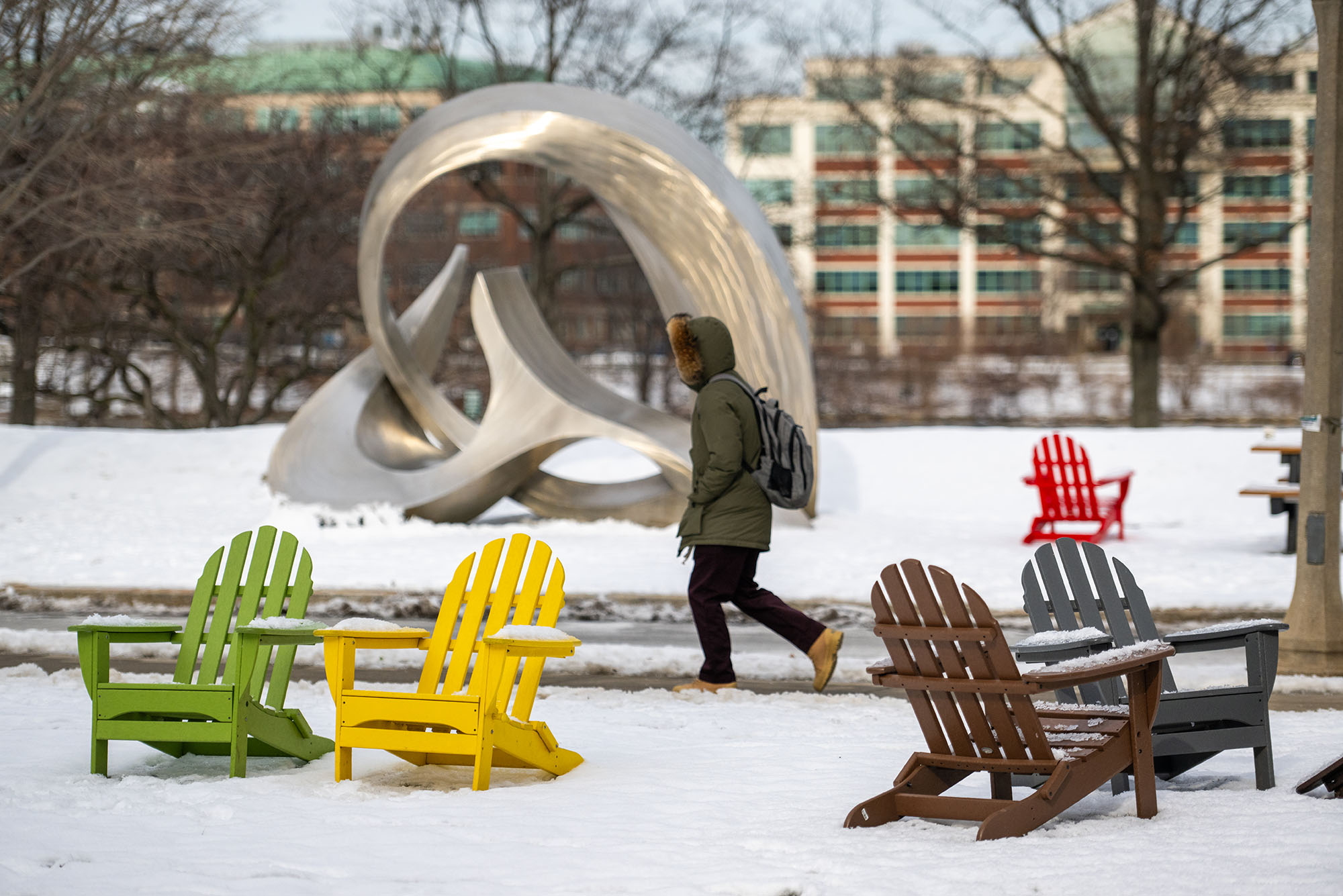 Photo: A student walks across a snow covered campus in January. A silver statue stands in the background and in the foreground, colorful chairs. The student is obscured by their winter coat.