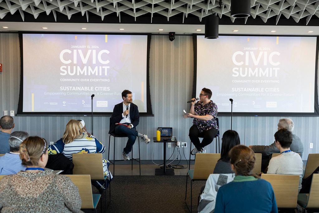 Photo: A fireside chat with Oliver Sellers-Garcia, left, Boston's first Green New Deal Director and a cabinet-level Senior Advisor to Mayor Michelle Wu, moderated by Jaye Meakem, the Boston Harbor Now's Community Engagement Manager. They sit on a stage with two projector screens behind them as they take questions from the crowd.