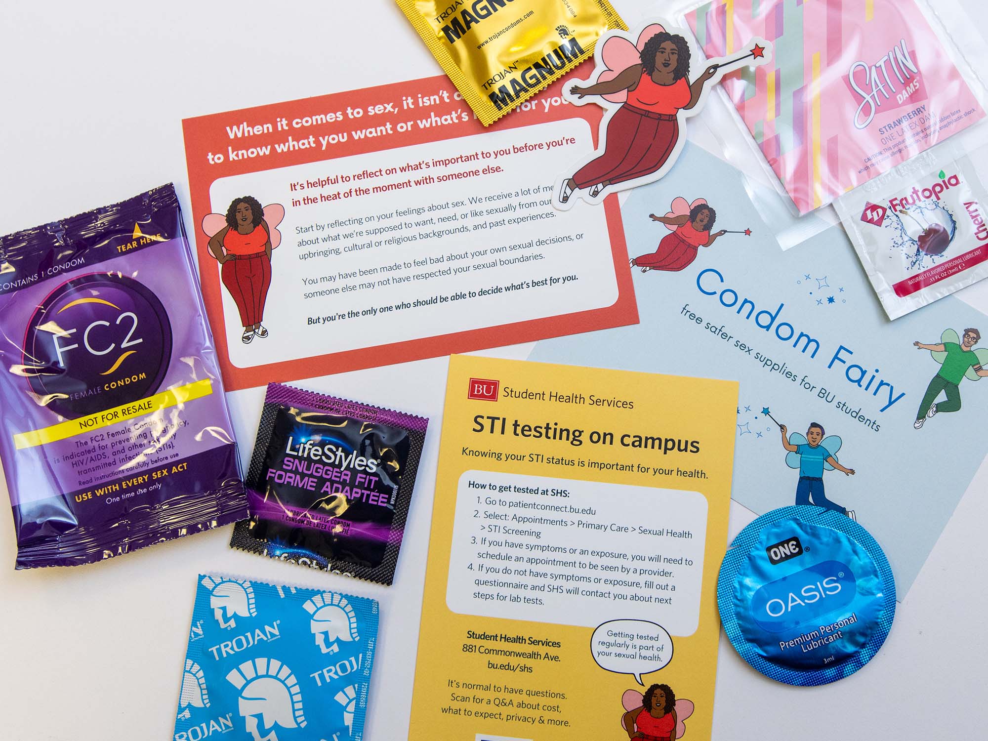Photo: A flat-lay of the supplies found when one orders from the Condom Fairy program located at BU. Various information cards and condoms are scattered on a white table. A featured image of the condom fairy characters in sticker form lay on top of the various cards.