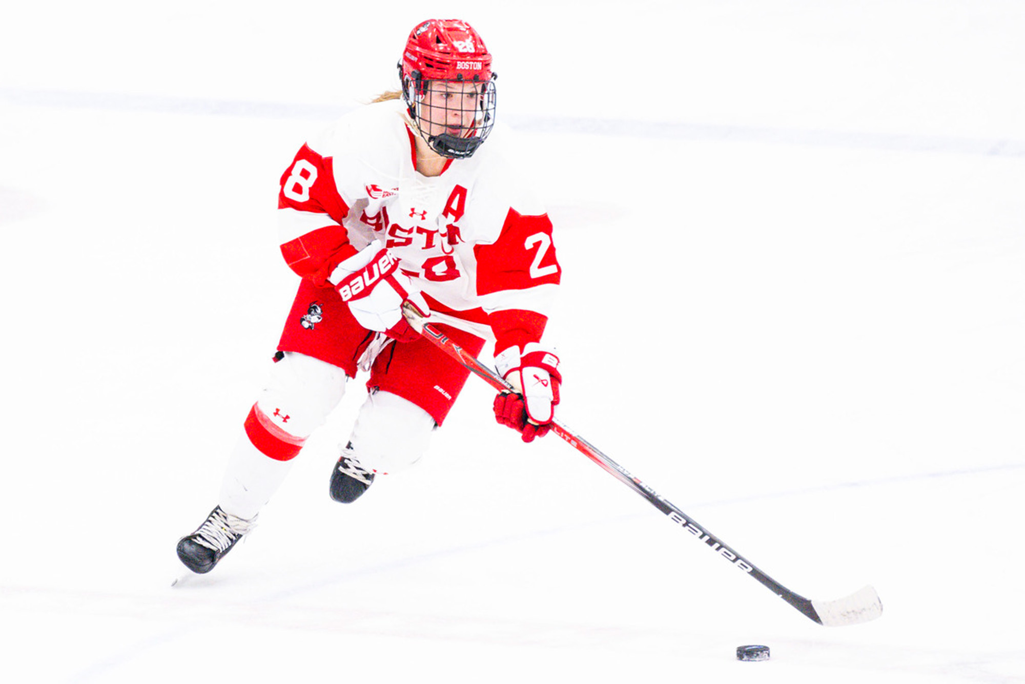 Photo: A BU women's hockey player in red and white uniform carries a puck down the ice during a recent game