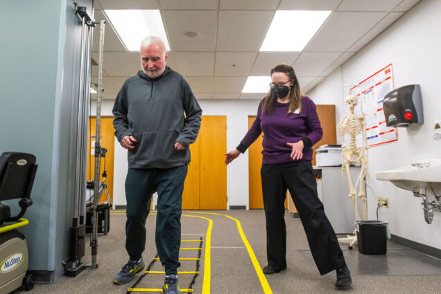 Photo: A man with white hair performance an exercise using a ladder on the ground, under the watchful eye of a PT next to him. The two appear to be in a clinic.