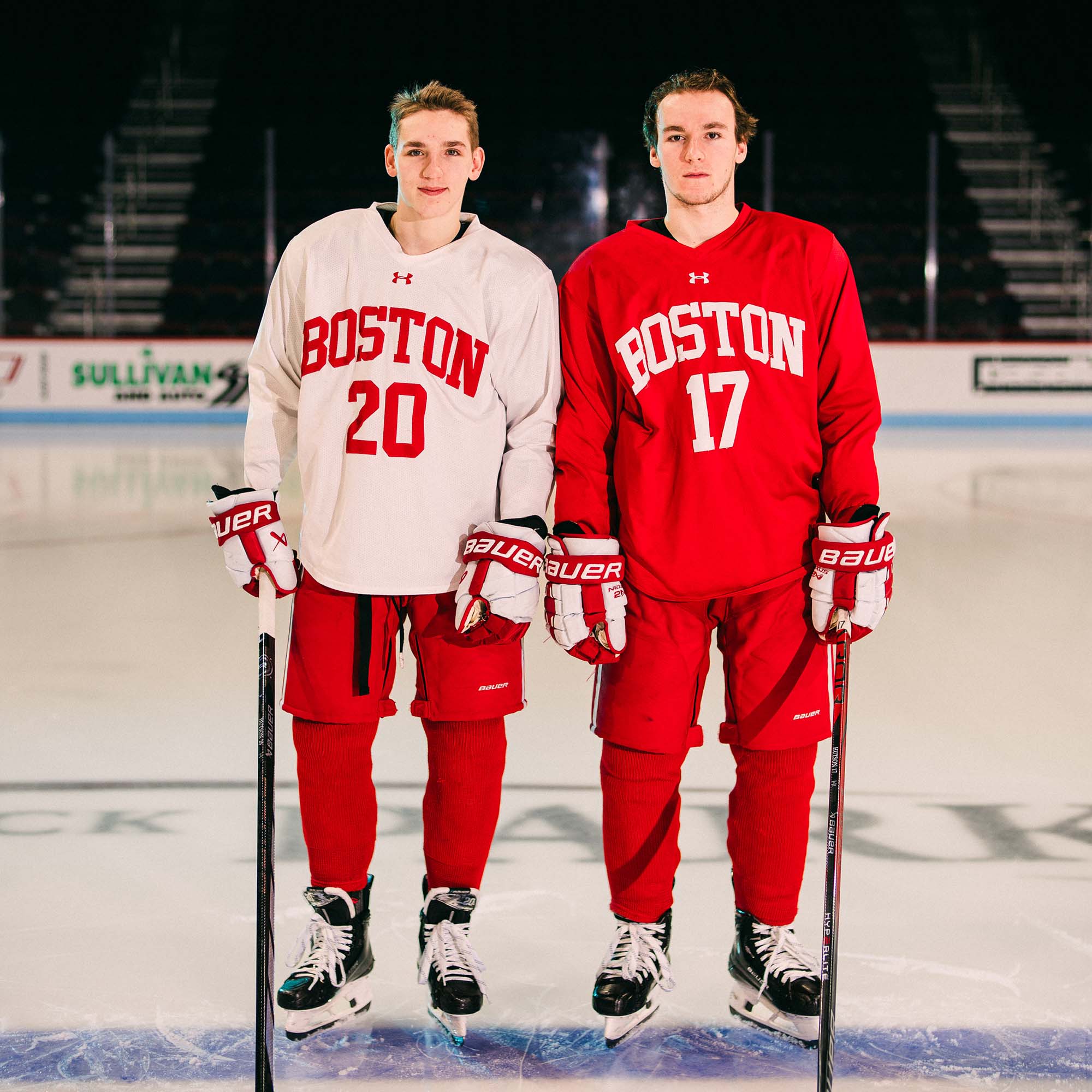 Photo: Two hockey players pose standing on the ice facing the camera. One wears a white practice jersey and the other, a red practice jersey and the red issue hockey pants.