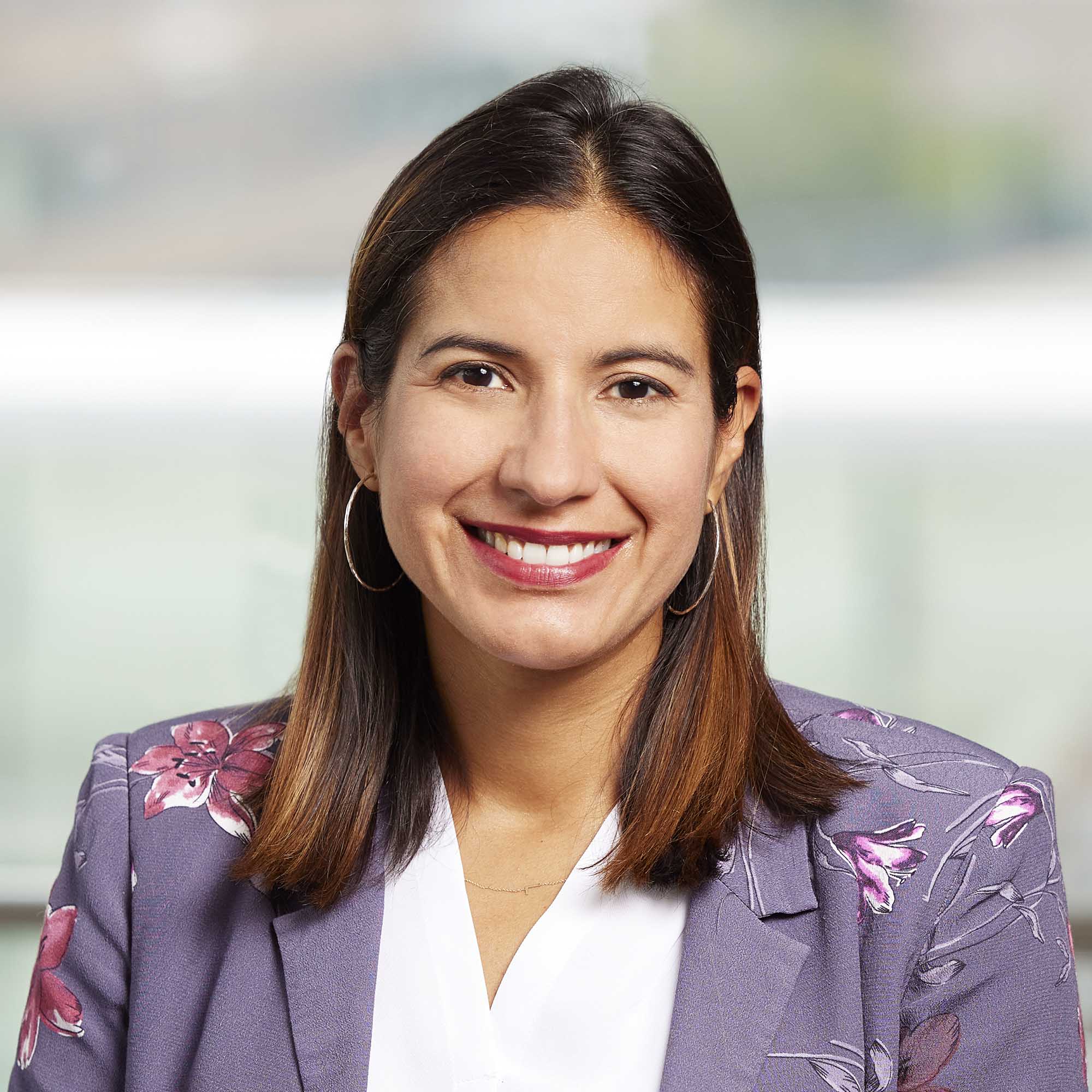 Photo: Ivania Rizo, a woman with shoulder length brown hair, smiles for a photo. She wears a purple blazer.