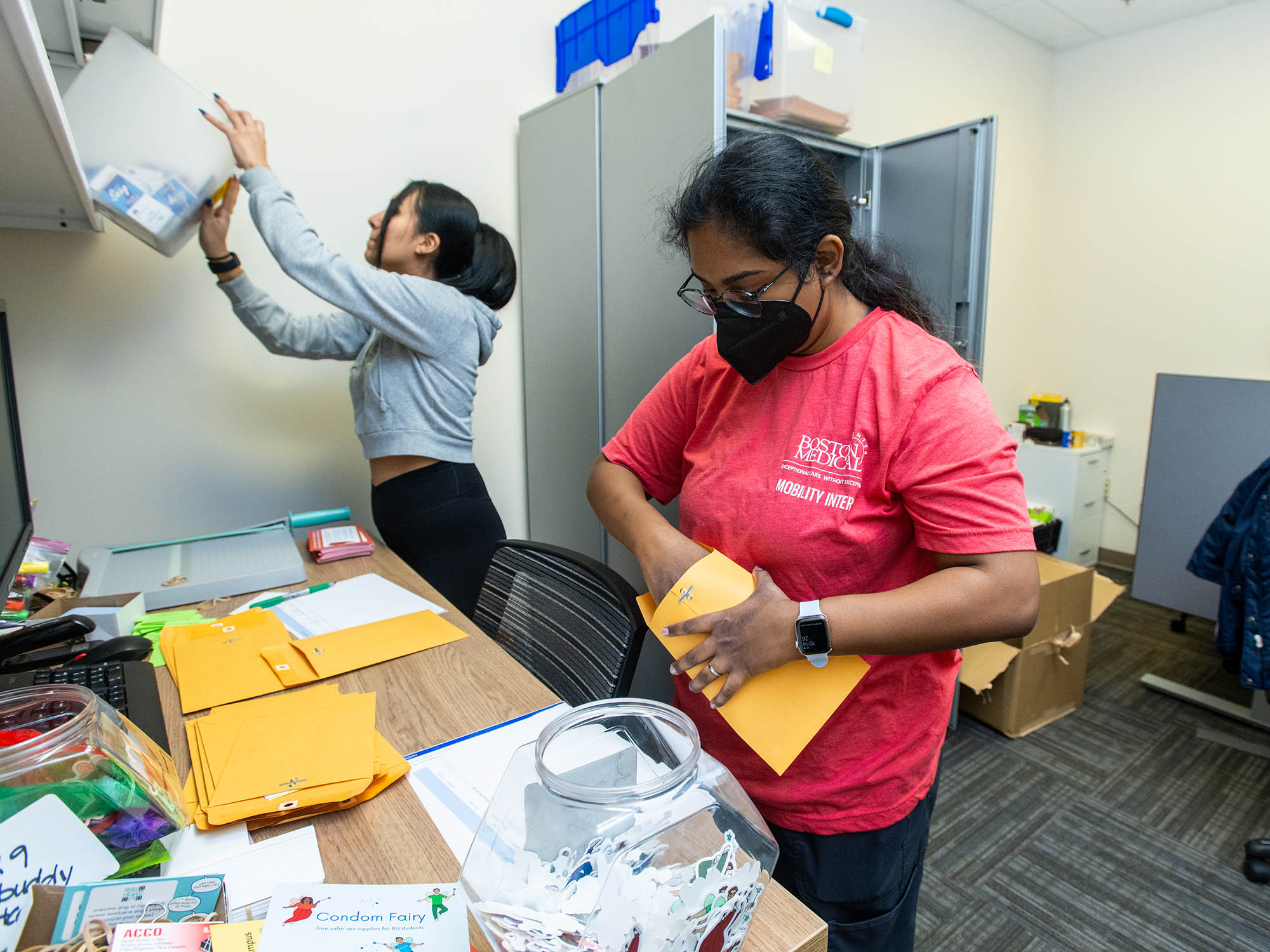 Photo: Two feminine-presenting individuals packing condoms. One, on the left background, wears a gray zip-up as they push a container in the cabinets. The front individual, wearing a red t-shirt, packs away the Condom Fairy materials into a mila envelope.