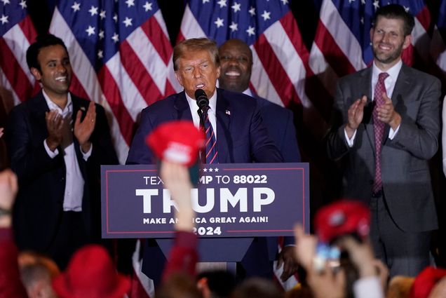 Photo: Republican presidential candidate former President Donald Trump speaks at a primary election night. He stands at a podium that reads TRUMP and an audience member reaches up to wave a red MAGA hat.