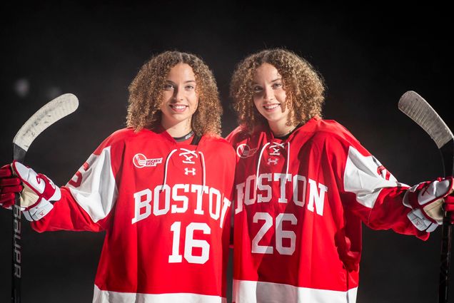 Photo: Two sisters with light brown curly hair pose in a studio for a shoot. They wear BU's hockey uniform--a red uniform with white lettering. They are both smiling.