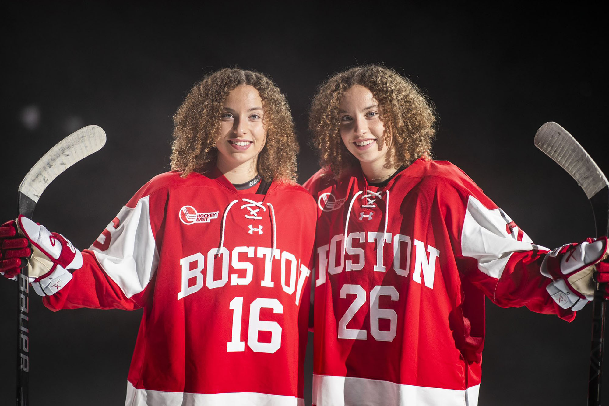 Photo: Two sisters with light brown curly hair pose in a studio for a shoot. They wear BU's hockey uniform--a red uniform with white lettering. They are both smiling.