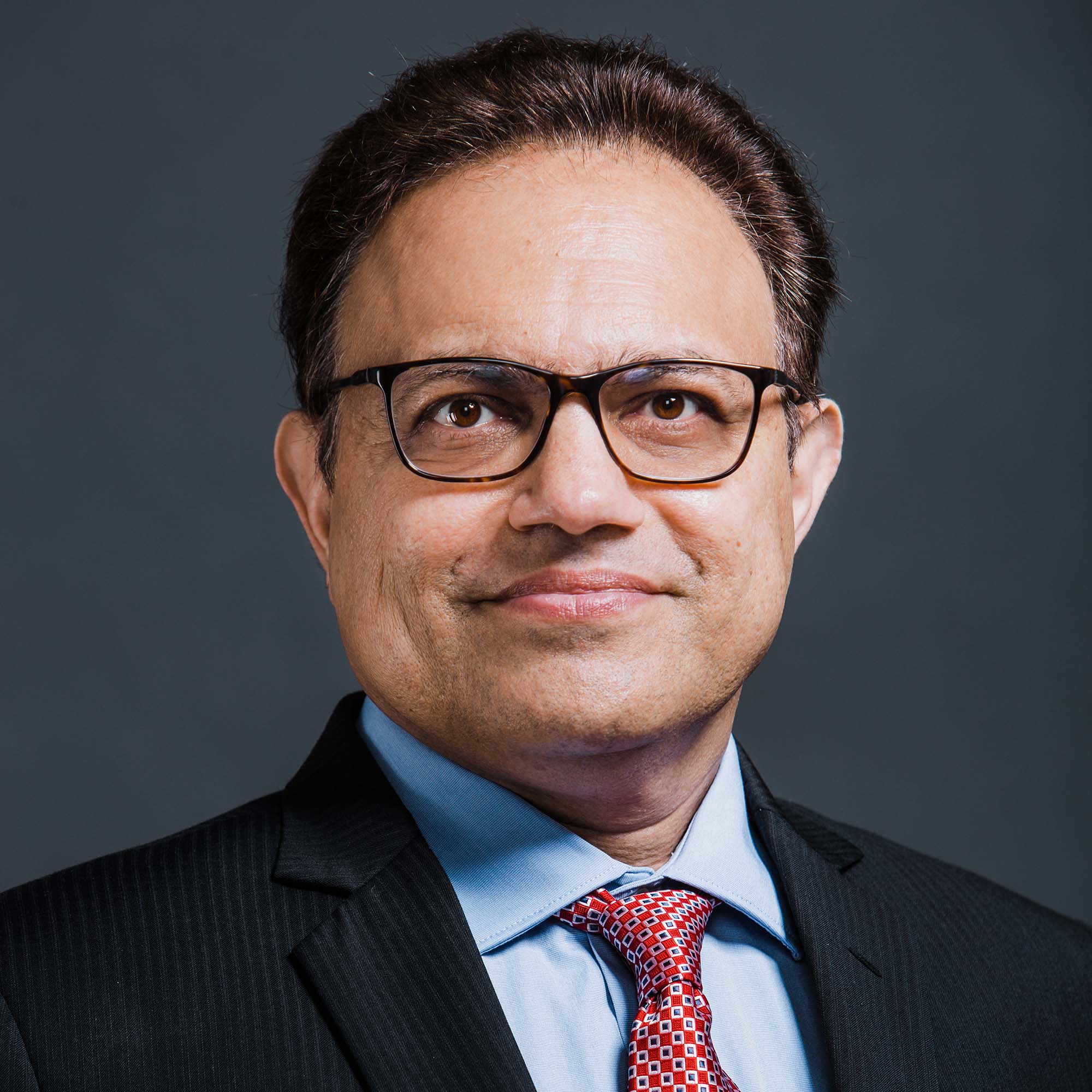 Photo: Arun Upneja, a Brown man and BU's SHA Dean, poses for a portrait photo. He wears a suit, a blue button up, and a red tie.