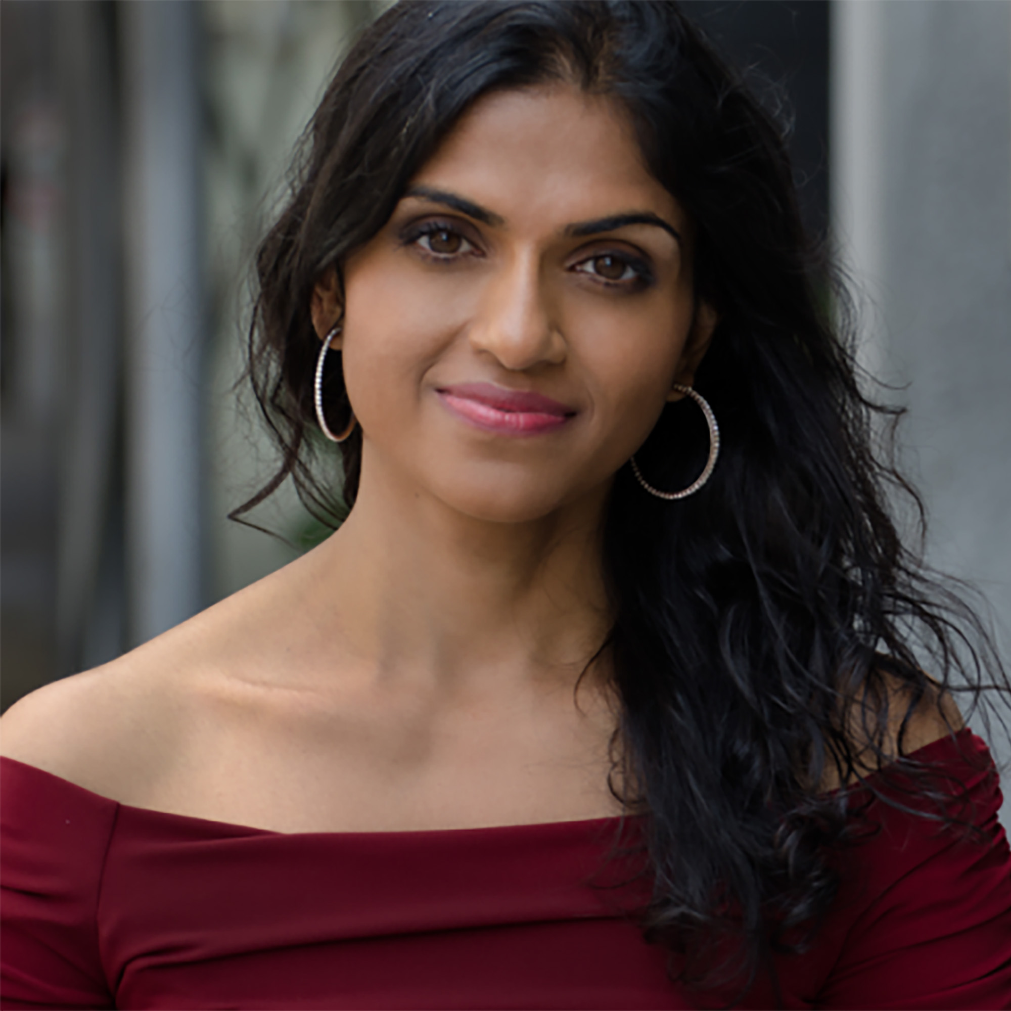 Photo: Saru Jayaraman, a Brown woman with long black hair, gives. a soft smile to the camera. She is wearing a maroon off-the-shoulder top and hoop earrings.