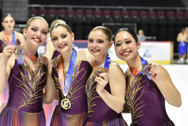 Photo: Four figure skaters hold up their medals after winning in a national championship. They smile big with purple uniforms.