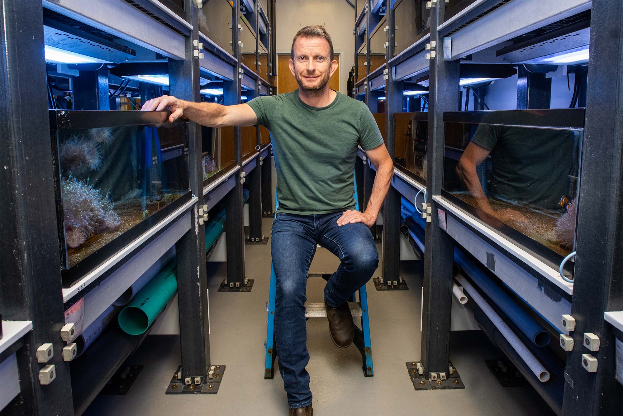 Photo: A man wearing jeans and a t shirt poses in a lab full of aquariums for clownfish
