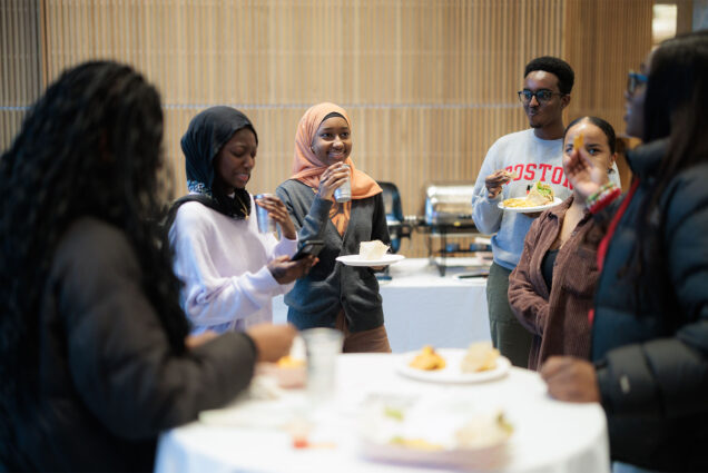 Photo: A group of college students gather around a table with appetizers and drinks. One of the is wearing a headscarf and another is wearing glasses.
