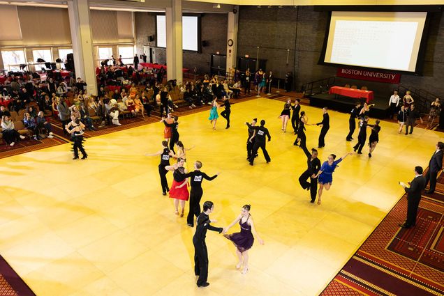 Photo: Multiple pairs of ballroom dancers dance their way across a large dancefloor in an open ballroom in front of a large crowd