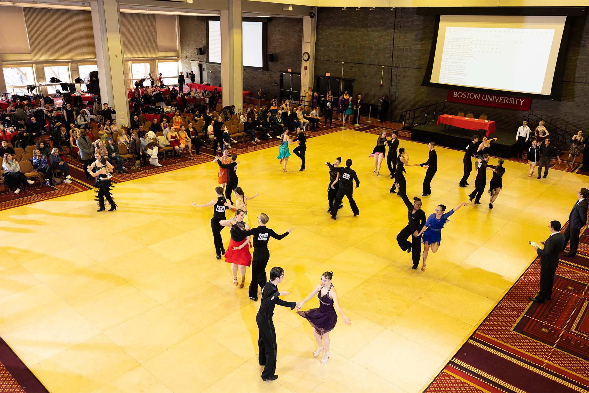 Photo: Multiple pairs of ballroom dancers dance their way across a large dancefloor in an open ballroom in front of a large crowd