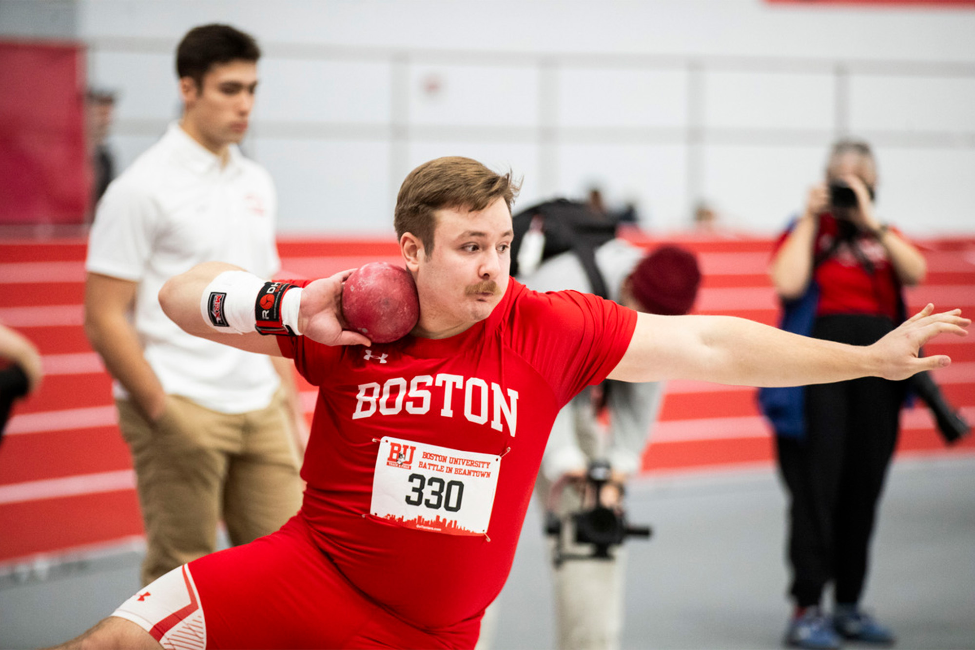 Photo: A college athlete wearing a red jersey throws a shotput at a track and field meet