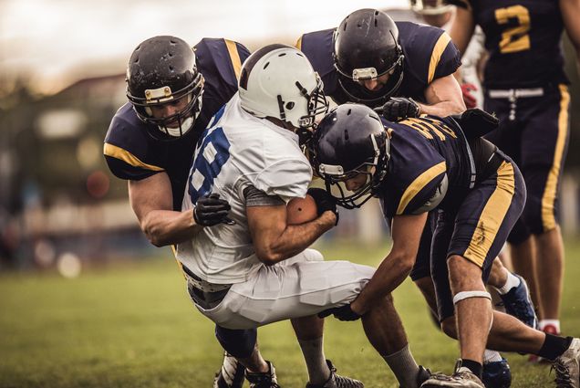 Photo: A group tackle of football players. One is wearing a white jersey and uniform and the others are in all black.