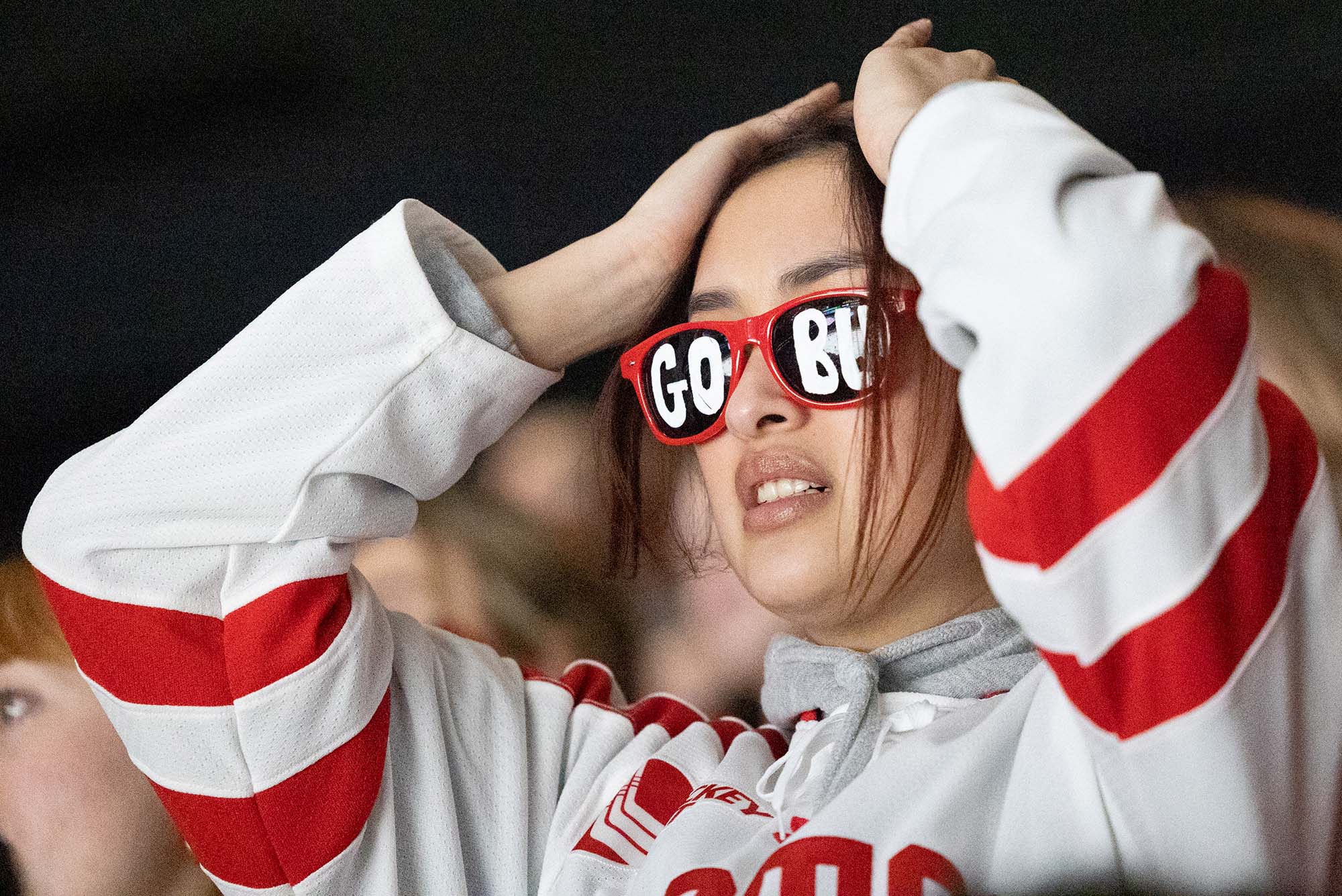 Photo: Fans react to the overtime game of college hockey for BU's Beanpot. A Asian woman wearing glasses that say GO BU holds her head in defeat.