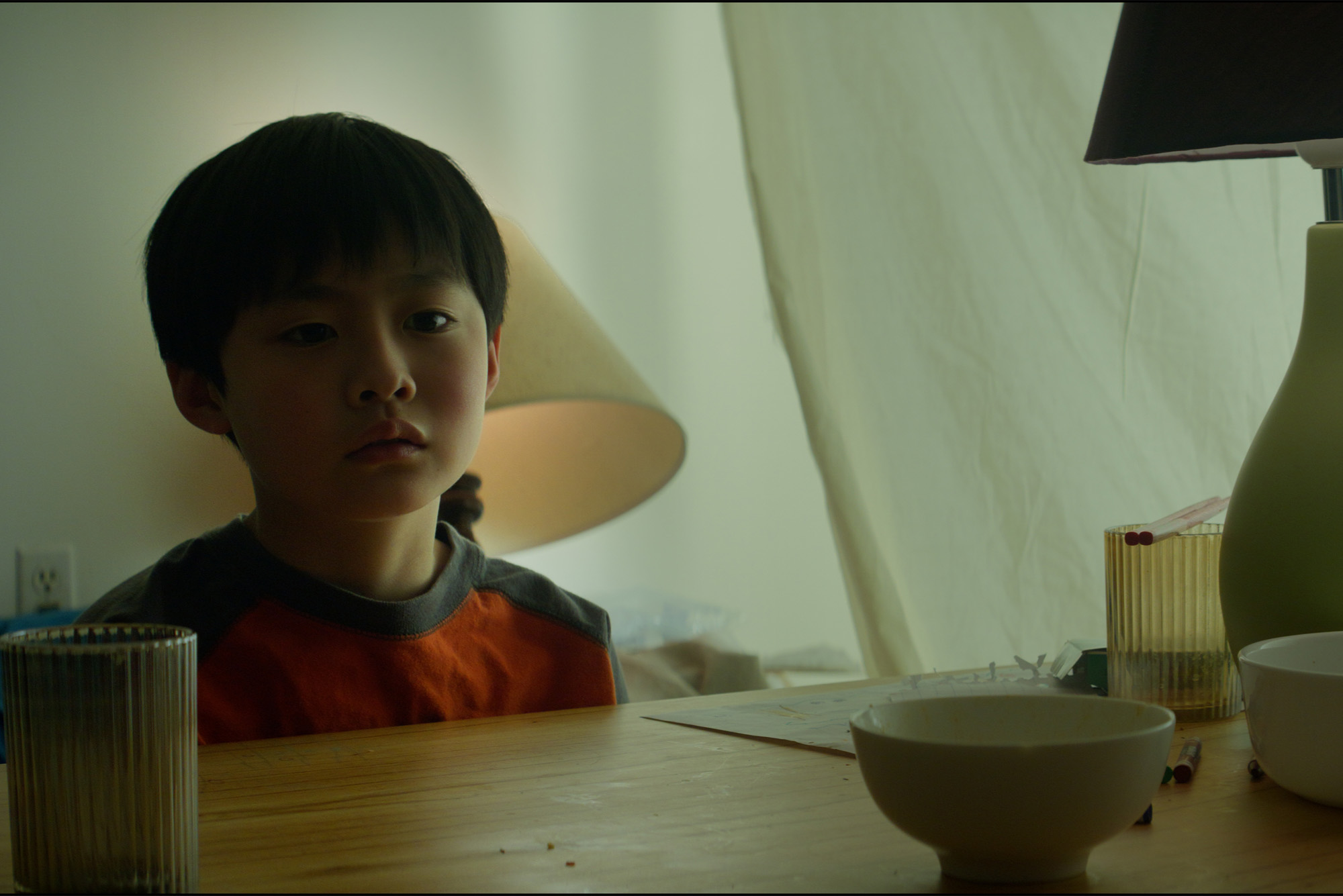 Photo: A young asian child stands in a dimly lit room near a lamp and a table with dishes on it