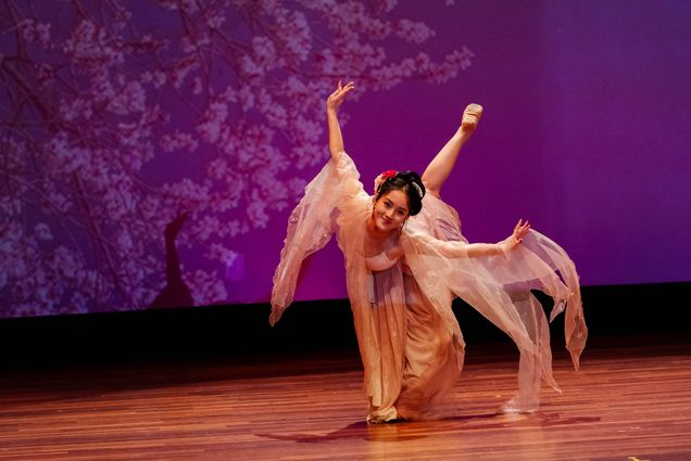 Photo: Yidi (Edie) Li, an Asian woman, performs on stage in a flowy, floral dress with her hair up in a fancy bun. The background behind her is purple with cherry blossoms projected at the back.