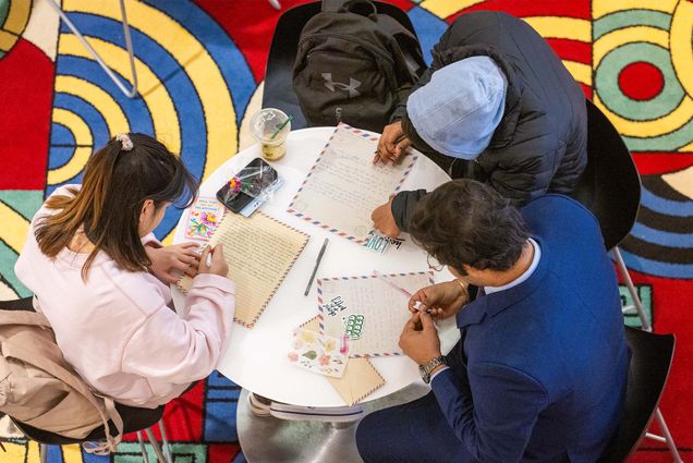 Photo: A group of students huddle around a table filled with writing utensils and notebooks. They are writing valentine's day cards for local senior centers.