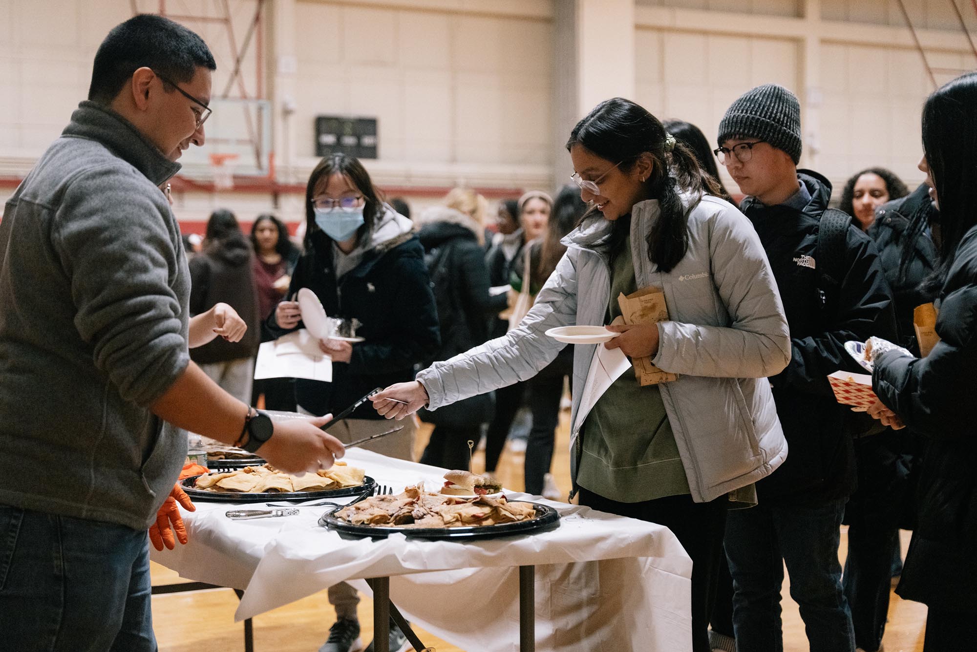 Photo: A young woman with a gray jacket reaches over the table to grab food that another person, a young man with a gray quarter zip, offers to her. They are both smiling.