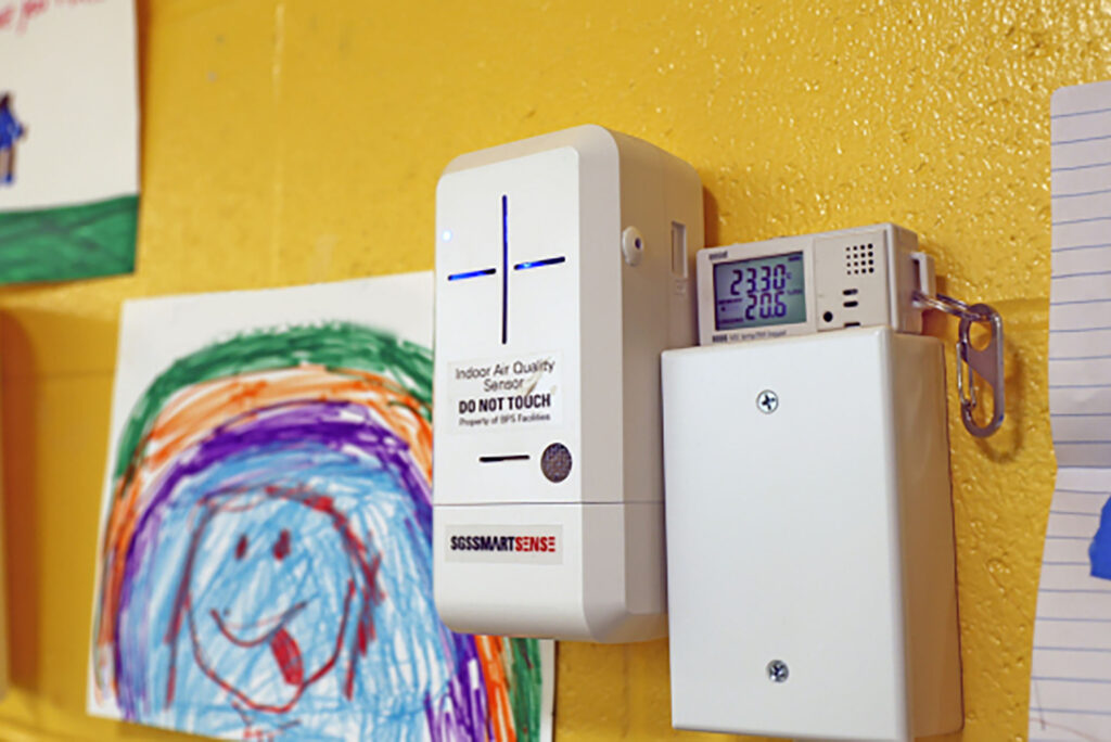 Photo: A white box with a cross of blue light on it. The device is an air sensor. It hangs on a yellow wall with children's drawing surrounding it.
