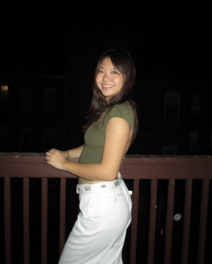 Boston University student Grace Kim. She is standing on a deck in the dark and the picture was taken with flash. She is turned to the side, her face smiling and looking at the camera. She has on a green top and white bottoms. 