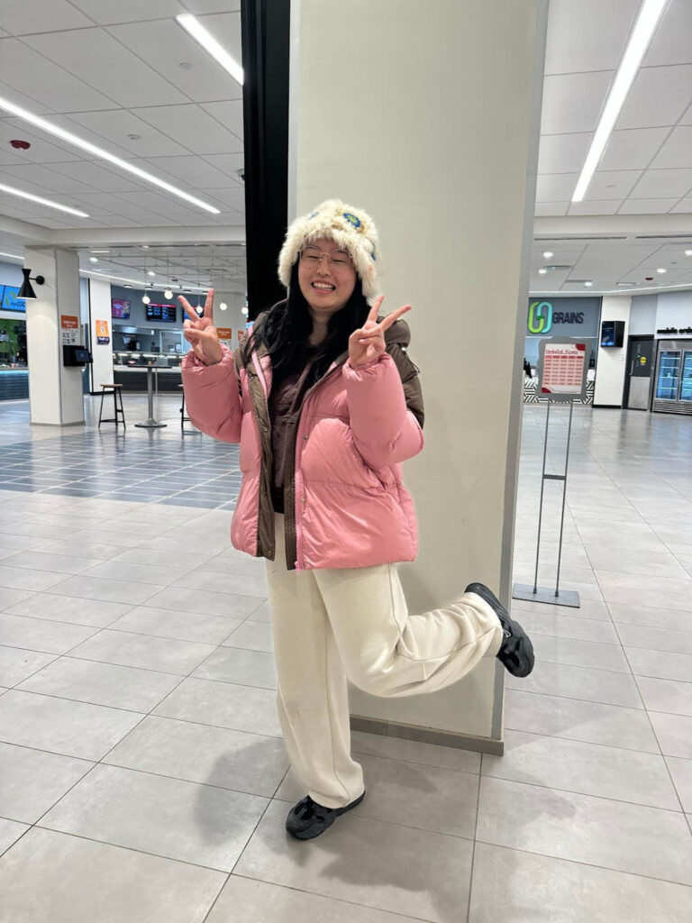 Photo: A photo of Wanjing Li, a young woman wearing black sneakers, a cream, wide-legged sweatpants, a baby pink puffer, and a fuzzy faux fur bucket hat.