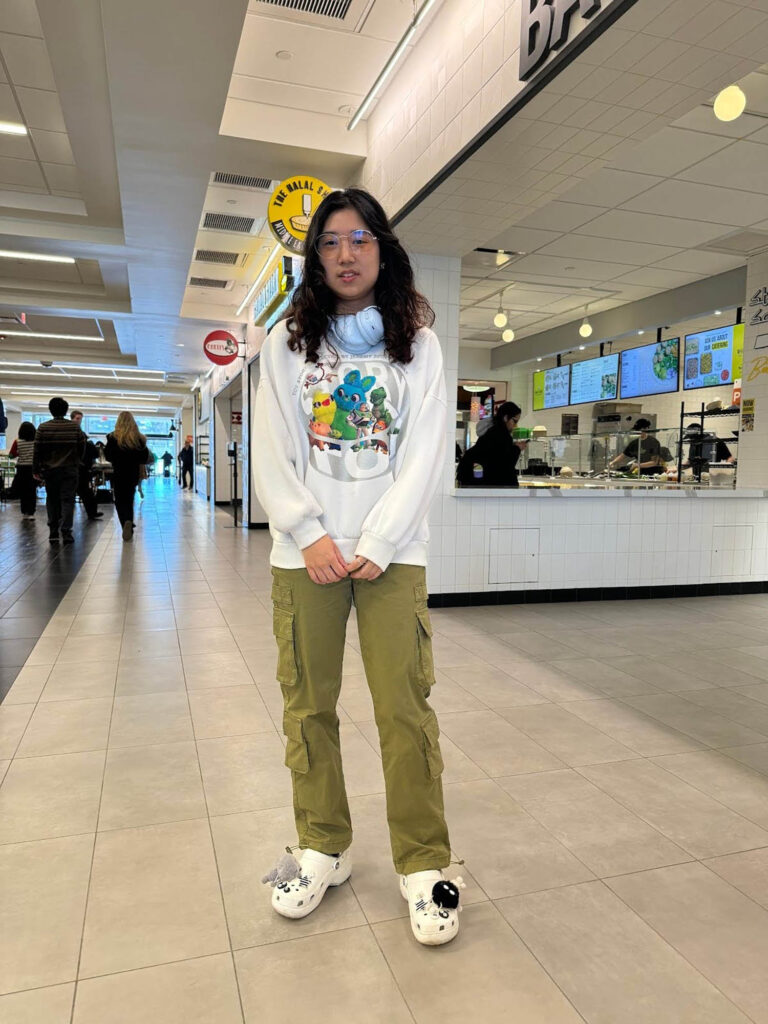 Photo: A photo of Willa Jiang, a young woman wearing platform crocs, green cargo pants, a thrifted sweatshirt with a unique plush design, and baby blue headphones around her neck.