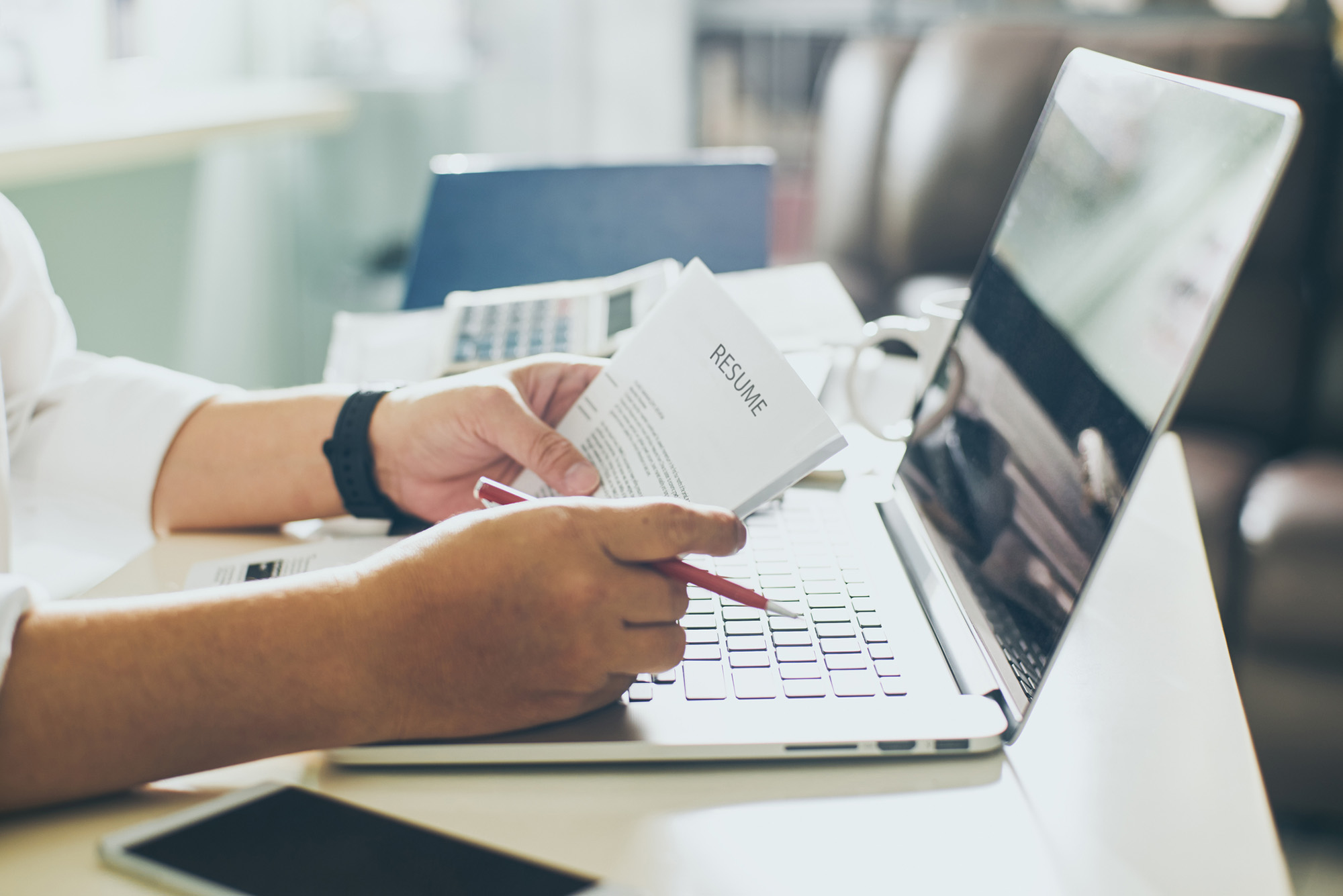 Photo: A stock image of someone looking through resumes with a laptop in front of them, pen in hand.