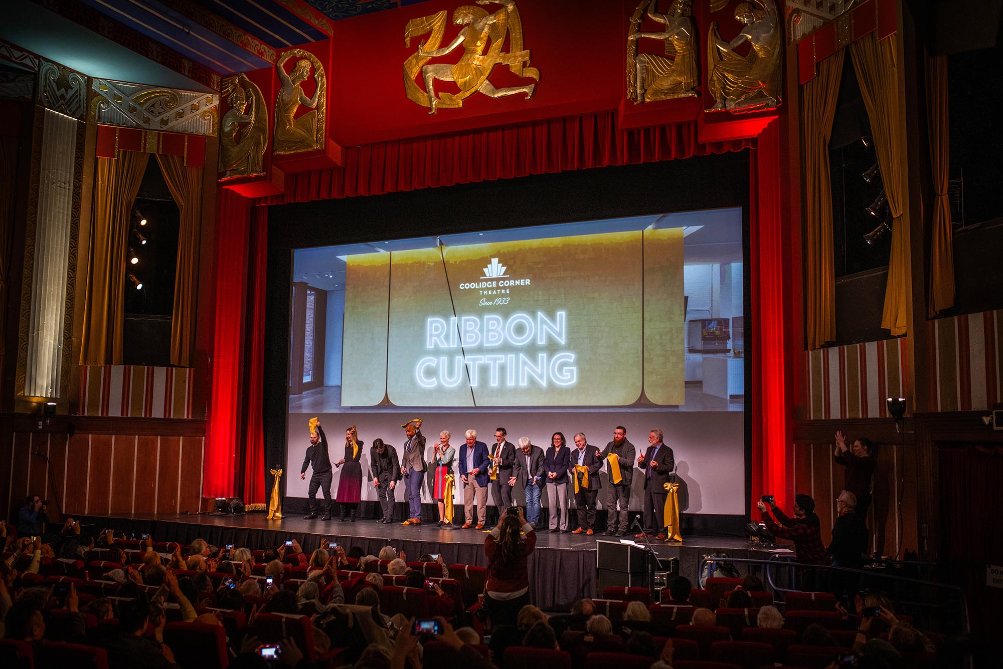 Photo: A picture of a stage with many people on it, all holding pieces of ribbon. There is a projection on the back of the stage that says "Coolidge Corner Theater, Since 1933, Ribbon Cutting"