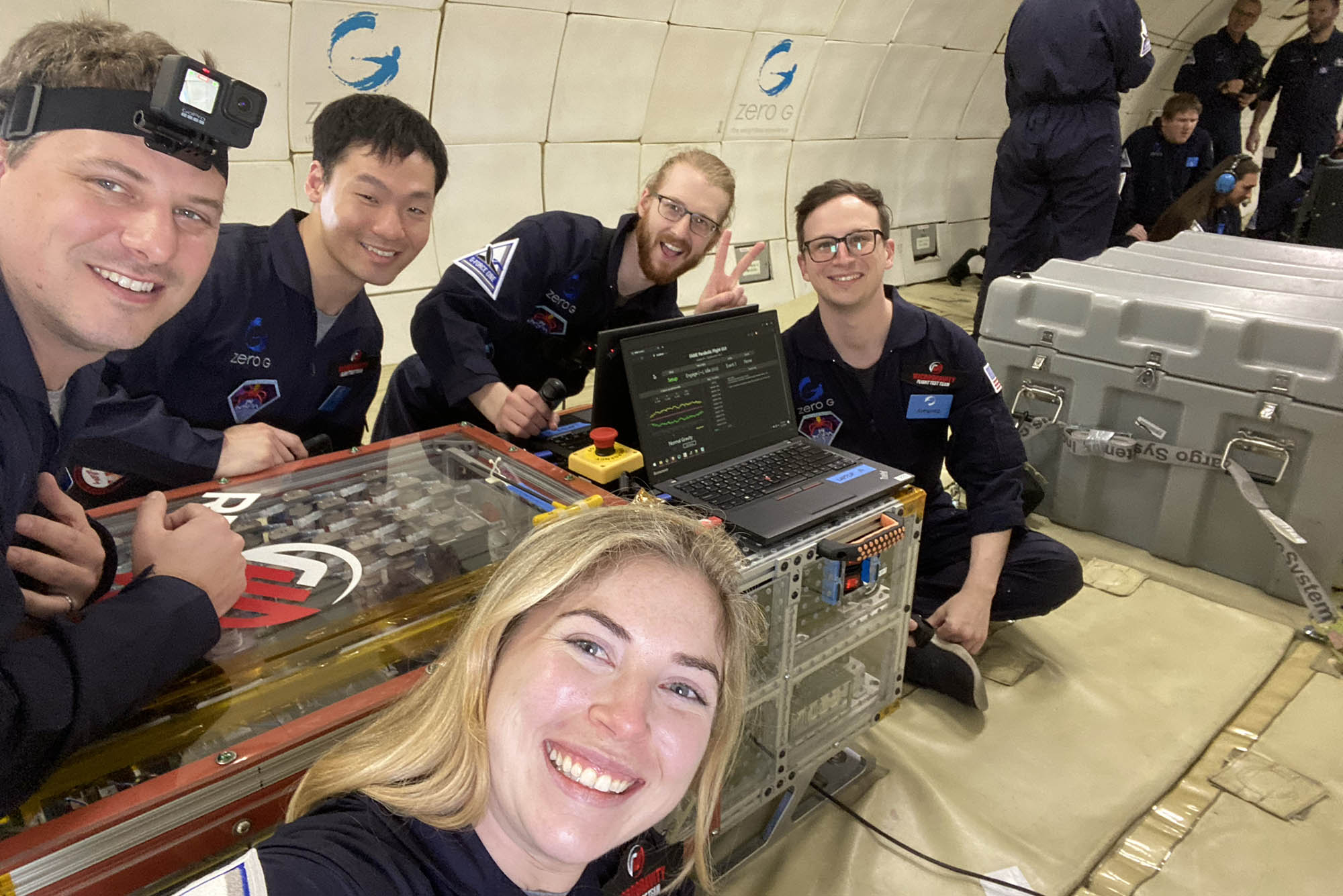 Photo: A group photo of Redwire Space Team in their navy jumpsuits. Four men in the back and one woman, Emma, in the foreground taking the photo.