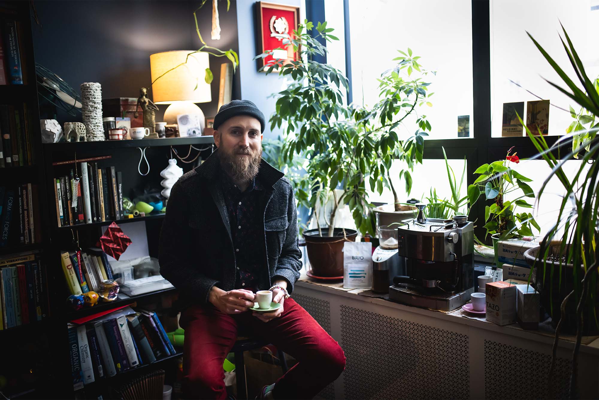 Photo: A man wearing a beanie and flannel sits in front of a window filled with plants, holding a small cup of coffee