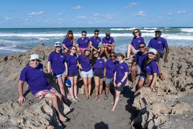 Photo: A group of volunteers in matching blue t shirts gather to pick up trash on a beach in Hobe Sound Florida