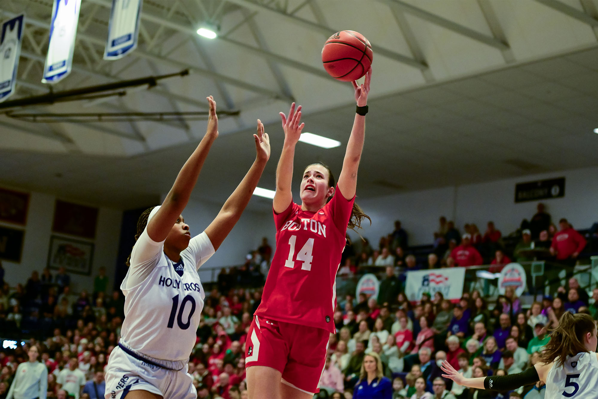 Photo: A Boston University women's basketball player shoots a layup during a recent game in a red jersey with white trim and the number 14. The layup is going over the outstretched arms of a defender in a white jersey with blue trim.