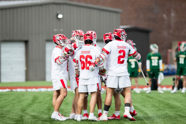 Photo: A group of BU college lacrosse players gather together in celebration after a goal. They are all wearing white jerseys with red numbers