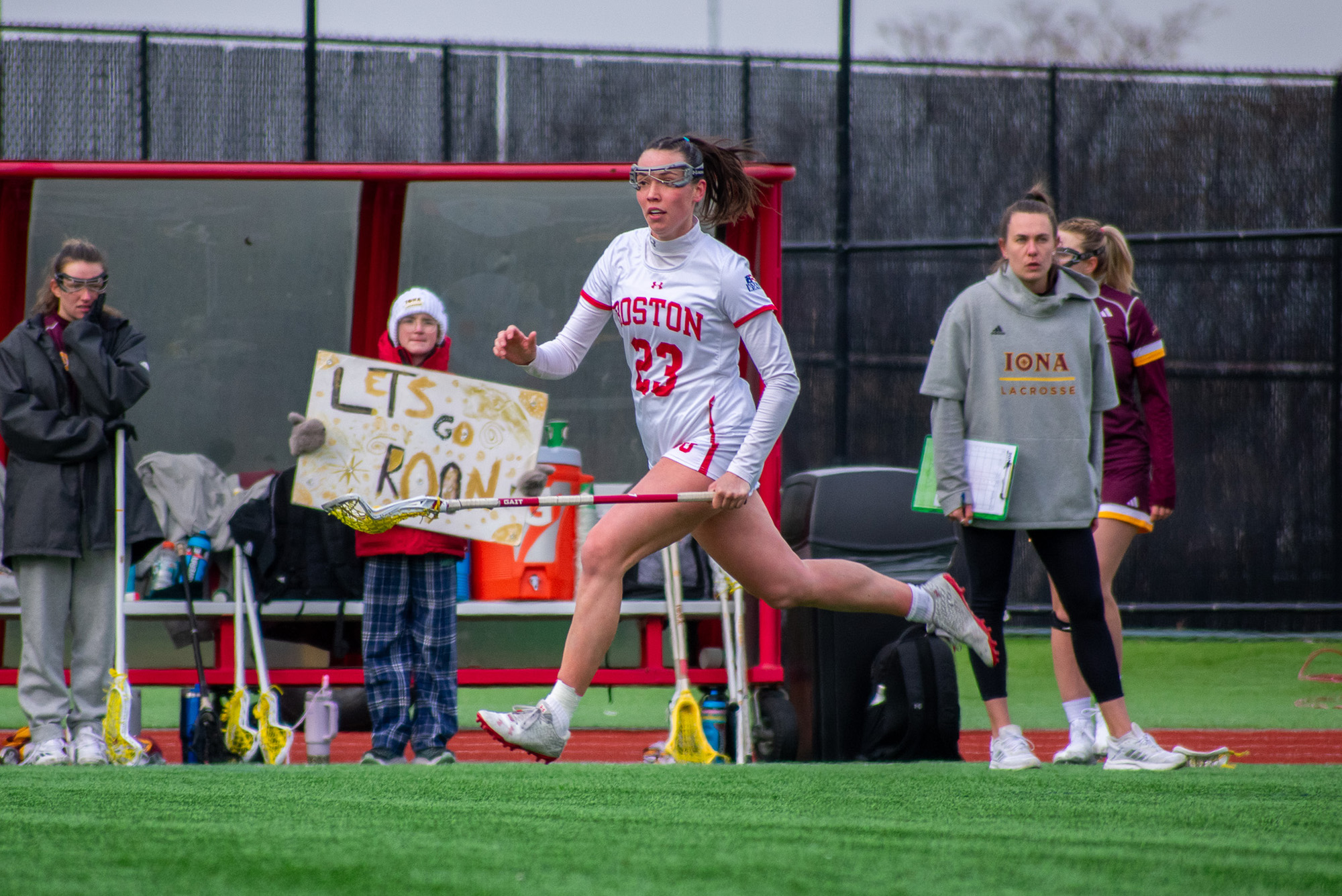 Photo: A woman's lacrosse player running up the sidelines. She is carrying a lacrosse stick and her uniform is white with red accents