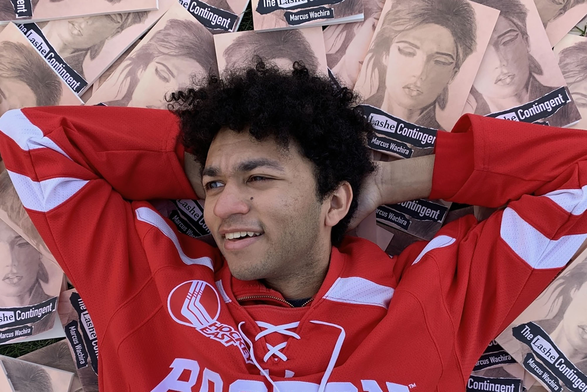 Photo: Marcus Wachira, a young man, with his hands behind his head and BU jersey laying on top of his artwork.