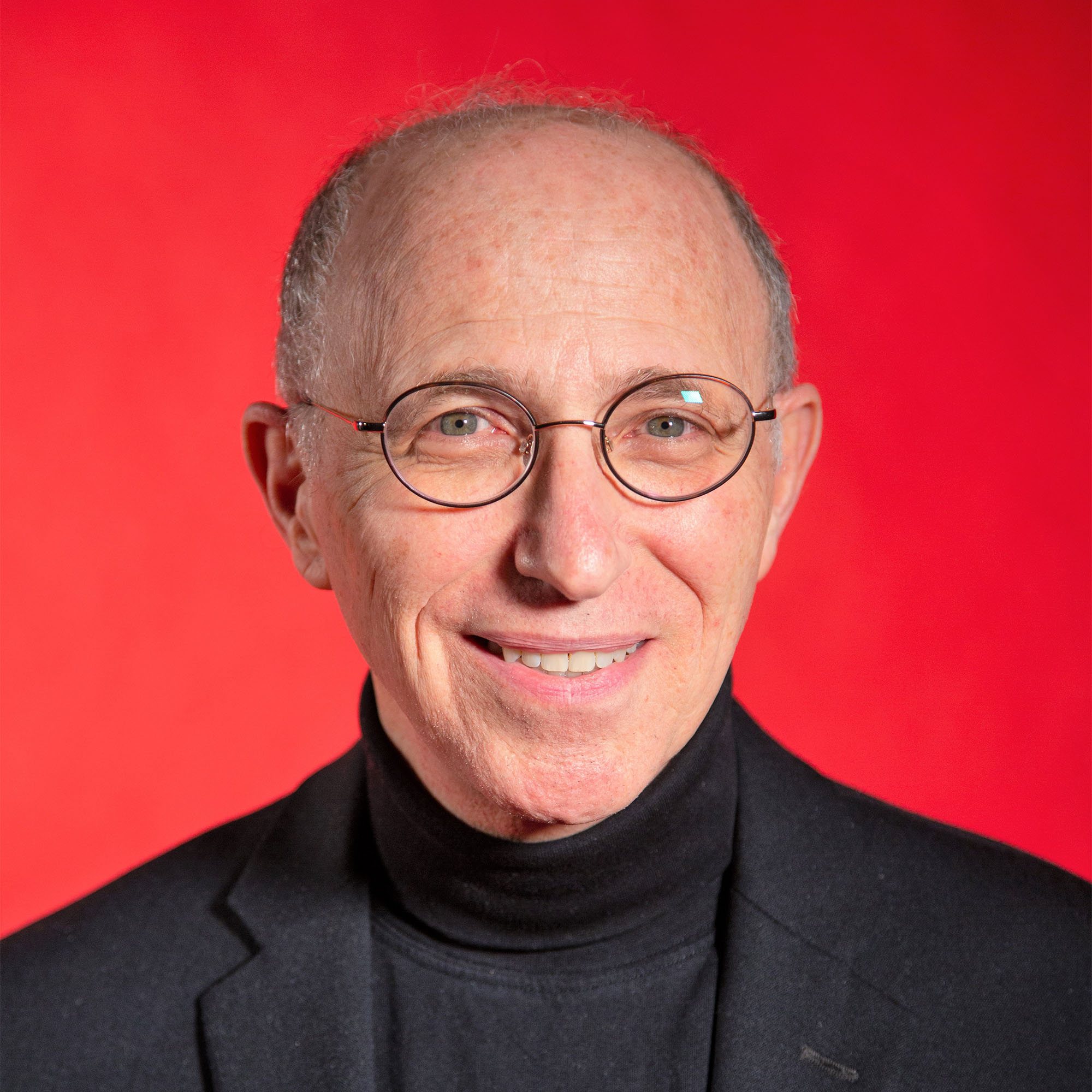 Photo: A middle aged white man wearing round glasses poses for a headshot in an elegant black suit and collared shirt in front of a red background