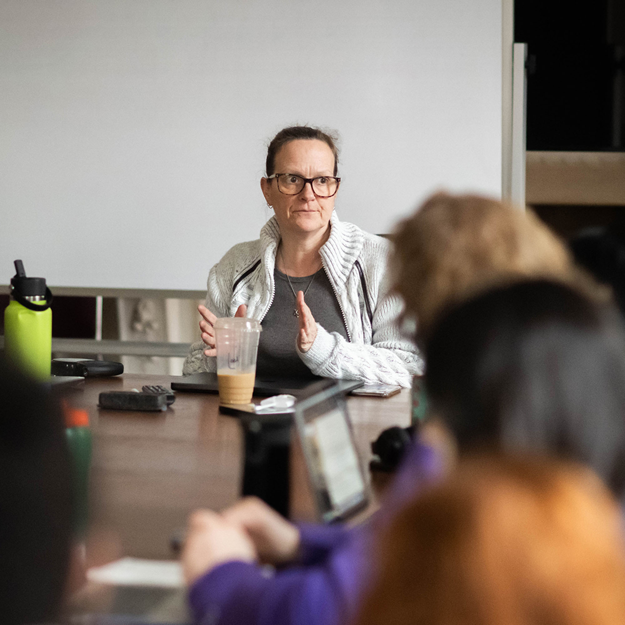 Photo: A classroom of people sitting around a large table. The teacher is talking and gesturing with her hands