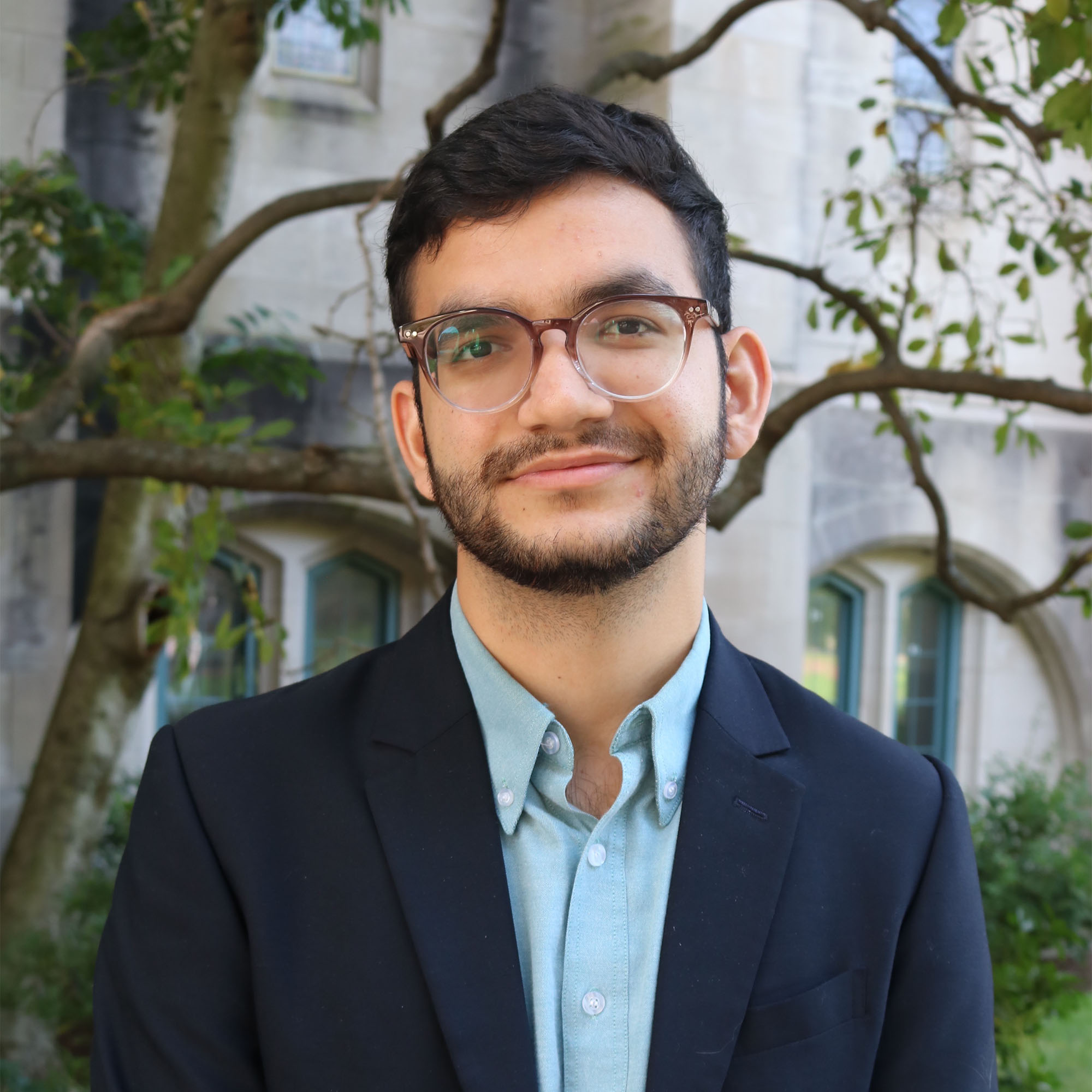 Photo: A man with dark hair and glasses wearing a blue collared shirt and suit jacket.