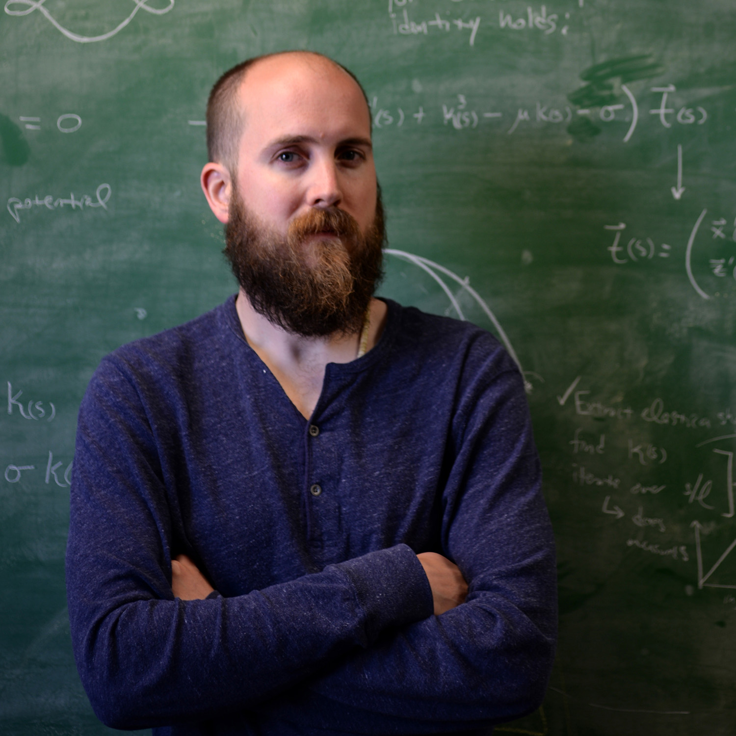 Photo: A picture of a man with a beard in a blue long-sleeved shirt crossing his arms. He is standing in front of a green chalk board that has white writing on it