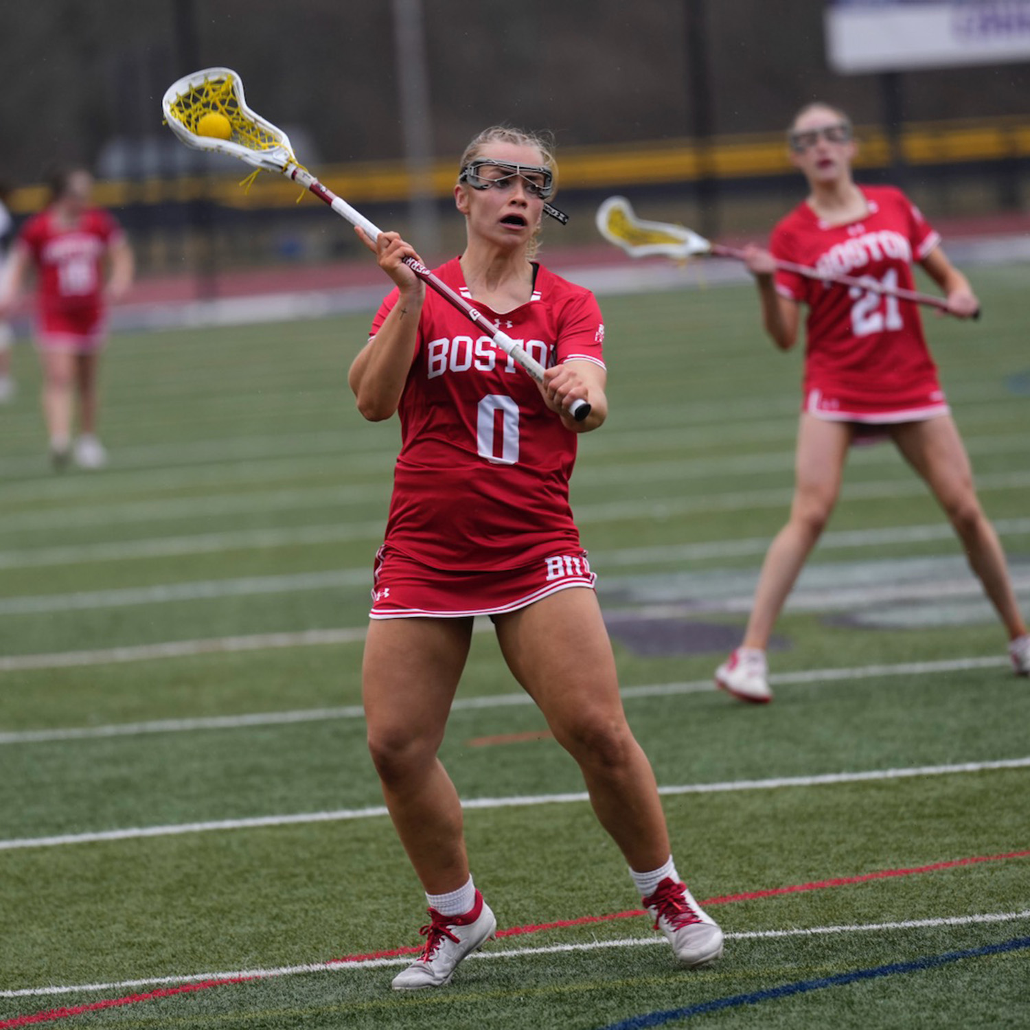 Photo: A woman's lacrosse defenseman looking to pass. She is holding a lacrosse stick and her uniform is red with white accents