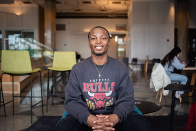 Photo: A young Black man sitting in a chair with a cross around his neck and a Chicago Bulls sweatshirt. He smiles for the photo.